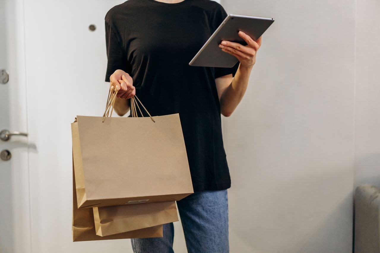 Woman in black shirt with shopping bags using a tablet at home, symbolizing online shopping convenience.
