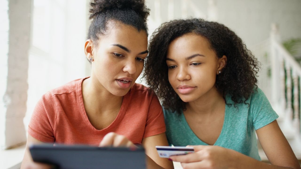 Two young women using a tablet and credit card while shopping online indoors.